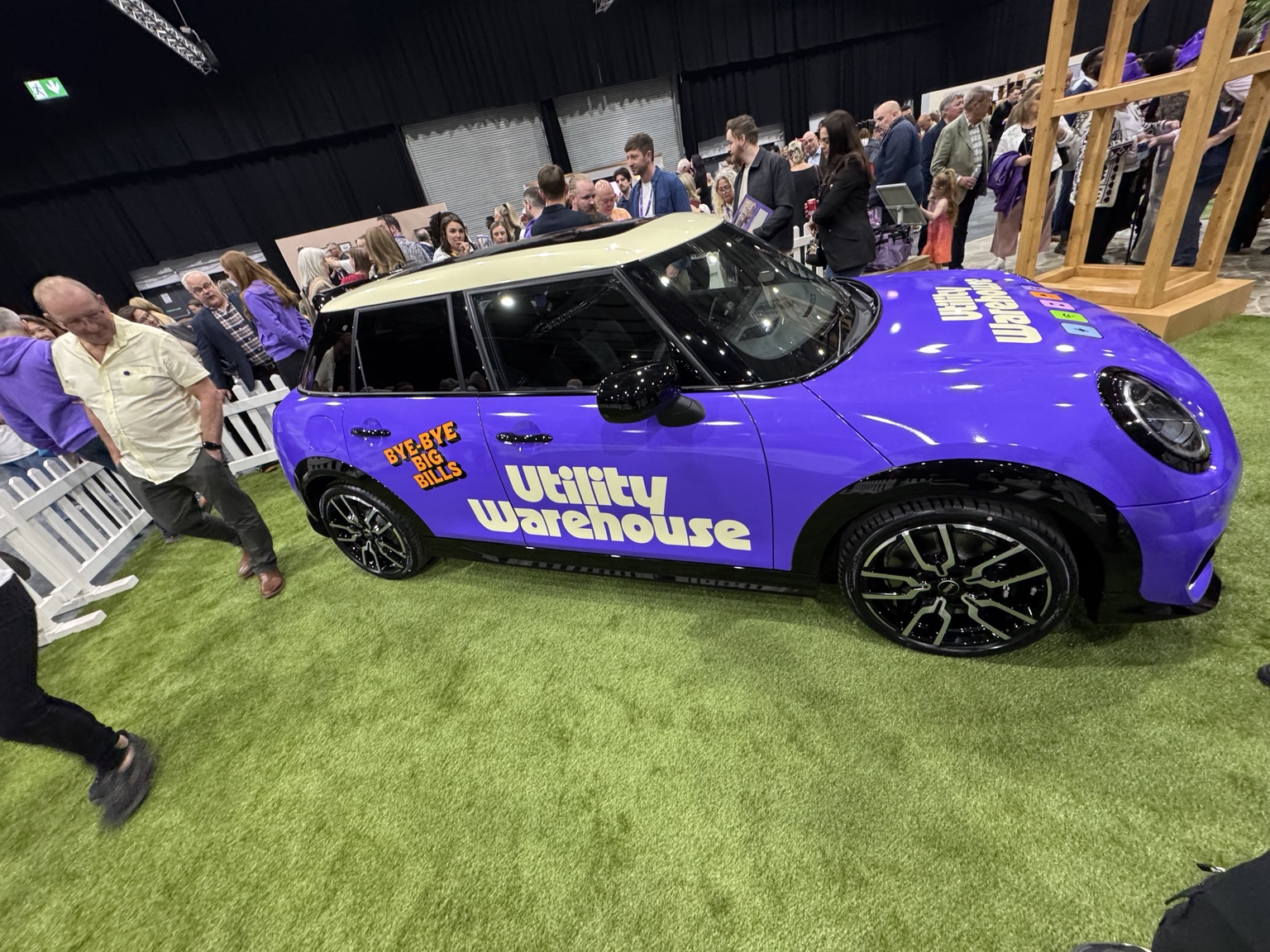 Side view of a purple MINI with a cream roof and Utility Warehouse lettering, on display on artificial grass at a busy UW event