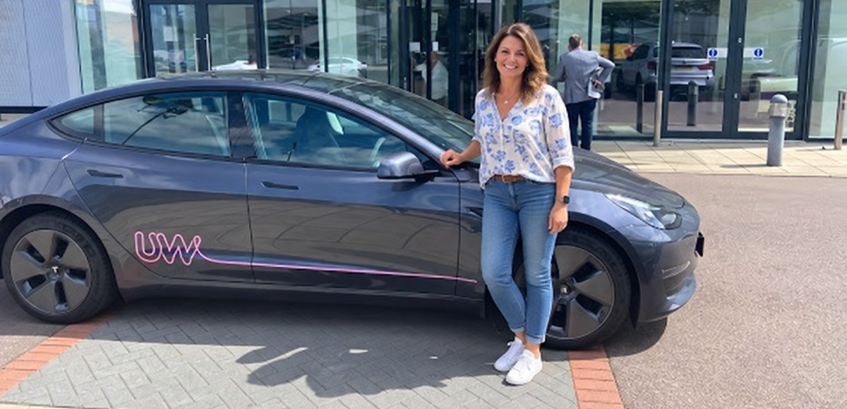A grey Tesla Model 3 with pink UW lettering parked outside the Utility Warehouse office entrance, with a woman standing beside it