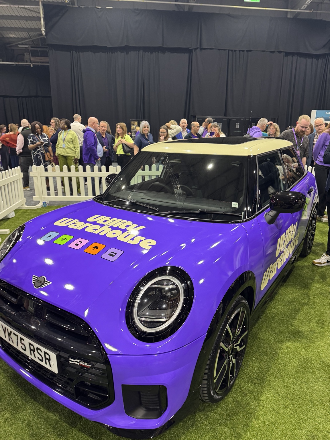 Front view of the purple Utility Warehouse MINI, with UW service icons on the bonnet and event guests behind it